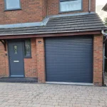 A grey roller garage door installed on a brick house with a grey composite doors, grey pvc windows upstairs and paved driveway.