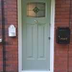 Green front door with vertical panels, a round door knocker, and a black letterbox on a brick wall.
