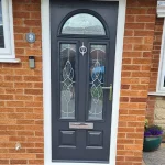 A dark gray contemporary composite front door flanked by dual narrow vertical windows, and a top arched window on a residential building. The door features a large silver handle, letterbox and big door knocker, white pvc framer and red brick left and right