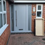 Grey composite front door with vertical panels and a glass sidelight. The door has a silver handle and letterbox.