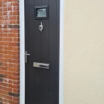 Black composite front door with vertical panels, a small rectangular glass panel, and a letterbox. The door has a silver handle and knocker.