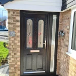 A black composite front door with a glazed panel and letterbox, flanked by brick walls and a sidelight window