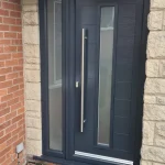 A dark gray contemporary composite front door flanked by narrow vertical windows on a residential building. The door features a large silver handle, a keyhole, and a kick plate. The entrance is framed by beige stone walls