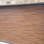 A wide garage door with horizontal wooden panels in a warm brown finish, set into a beige stone building. The door's design mimics natural wood grain, and it's framed on one side by a rough stone wall with a traditional lantern mounted above