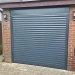 Grey Roller Garage Door, with gutter and roof slates graduating upwards, red brick wall on the left, and extended brick on the right hand side with a small pvc window, a block paving driveway