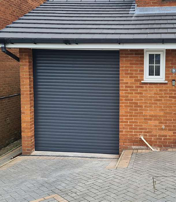 Grey Roller Garage Door, with gutter and roof slates graduating upwards, red brick wall on the left, and extended brick on the right hand side with a small pvc window, a block paving driveway