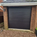 Roller Garage Door, with gutter and roof slates graduating upwards, pebbled driveway, and brick either side of the entrance