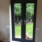 View of a garden through an open double-glazed French door with black frames, showing a lush green lawn and trees. The interior walls around the door are partially unfinished with visible plaster and electrical sockets.