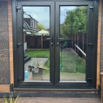 Modern black-framed French doors on a wooden exterior wall, opening to a backyard garden with green grass and outdoor furniture.