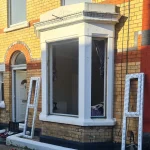 A brick house with a bay window under construction. New white window frames are leaning against the wall.