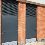 Two identical dark gray metal doors set in separate sections of a red brick wall, each featuring a handle, keyhole, and a blue sticker. Above each door is a large black metal ventilation grate. The base of each doorway has a protective blue and white waterproof membrane visible