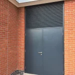 A dark gray metal door set within a red brick wall, featuring a single handle and a keyhole. Above the door is a large metal grate for ventilation, and the door is surrounded by a light gray concrete ledge and small pebbles at the base