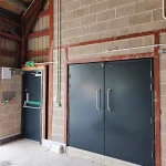 A set of large, dark gray double doors with horizontal handles on a block wall inside a building with exposed wooden beams and metal pipes. To the left, there is a smaller single door with a green 'Push bar to open' sign and a fire exit sign above it
