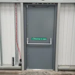 A grey fire exit door equipped with a horizontal push bar and safety signage. Signs include a green 'Fire exit' sign with a running figure icon and a 'Push bar to open' sign with a checkmark
