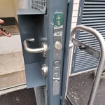 Close-up of a gray metal door's locking mechanism, showing multiple security bolts and two metal handles. The door is slightly ajar, and part of a turnstile with a hand pushing it is visible to the right