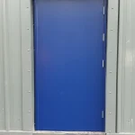 A bright blue door set within a grey metal frame, featuring small white hinges on the right side, mounted on a textured grey wall. The ground in front of the door is partially visible, showing a black asphalt surface