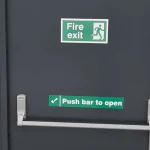 A grey fire exit door equipped with a horizontal push bar and safety signage. Signs include a green 'Fire exit' sign with a running figure icon and a 'Push bar to open' sign with a checkmark